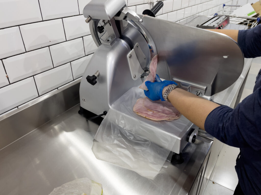 Image of hands of a deli assistant wearing disposable plastic gloves using ham slicer machine.