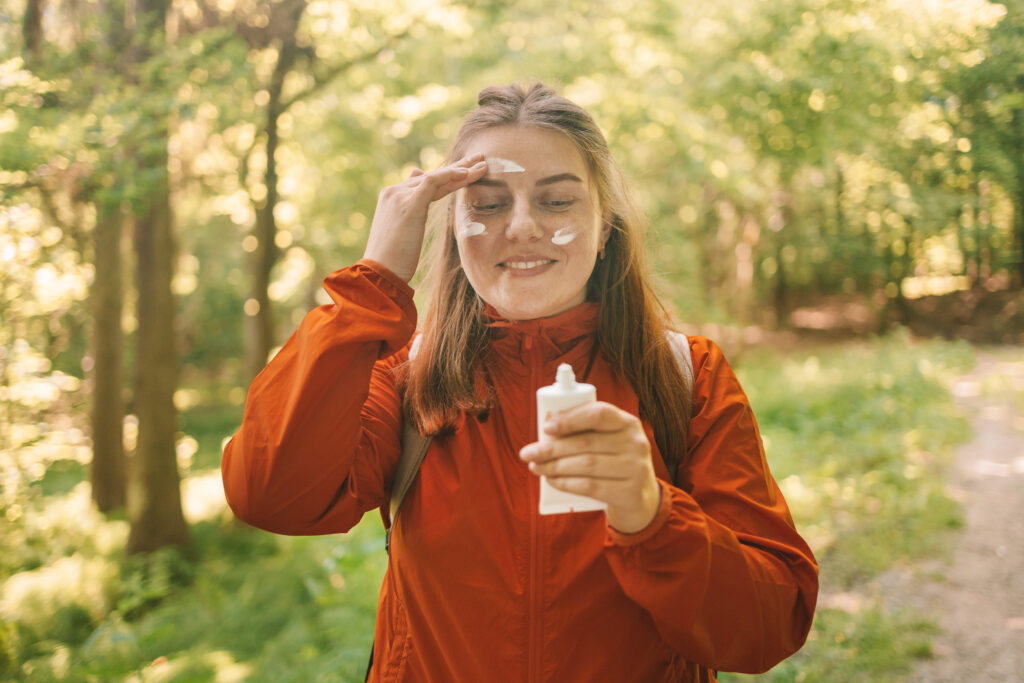 A young woman in a long-sleeved orange sport jacket applying sunscreen while hiking in the mountains. 